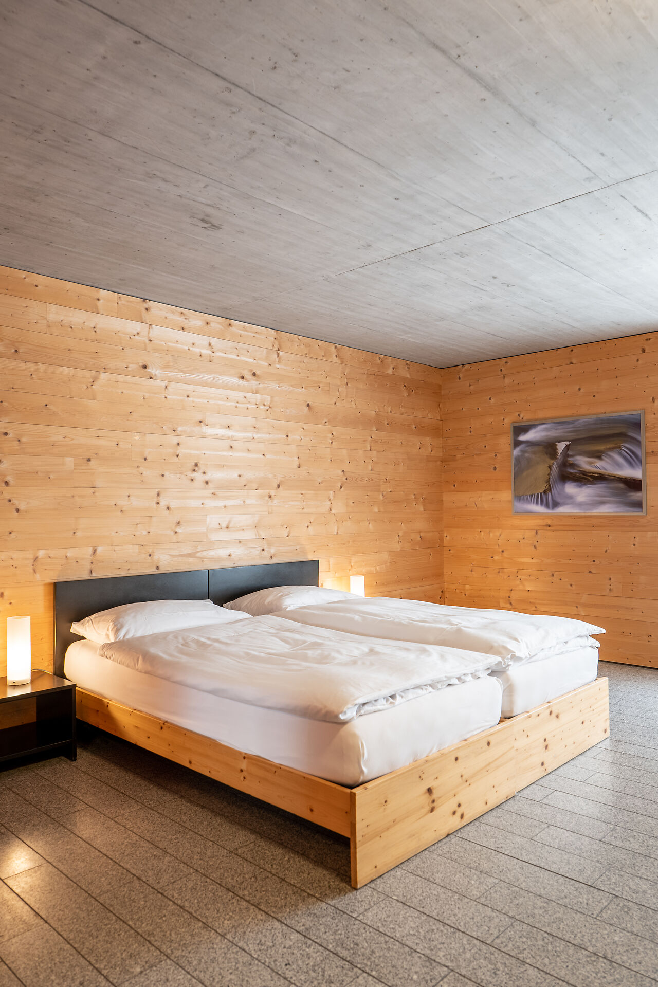 Wohn- und Schlafbereich eines Studio S der Inn Lodge mit einem Doppelbett mit weisser Bettwäsche und einem schwarzen Nachtisch.  An der grauen Wand hängt ein gerahmtes Bild. Die holzverkleidete Wand und der Holzboden prägen den Raum.