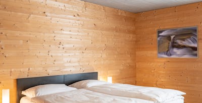 Studio S | ALL IN ONE HOTEL INN LODGE Wohn- und Schlafbereich eines Studio S der Inn Lodge mit einem Doppelbett mit weisser Bettwäsche und einem schwarzen Nachtisch. An der grauen Wand hängt ein gerahmtes Bild. Die holzverkleidete Wand und der Holzboden prägen den Raum.