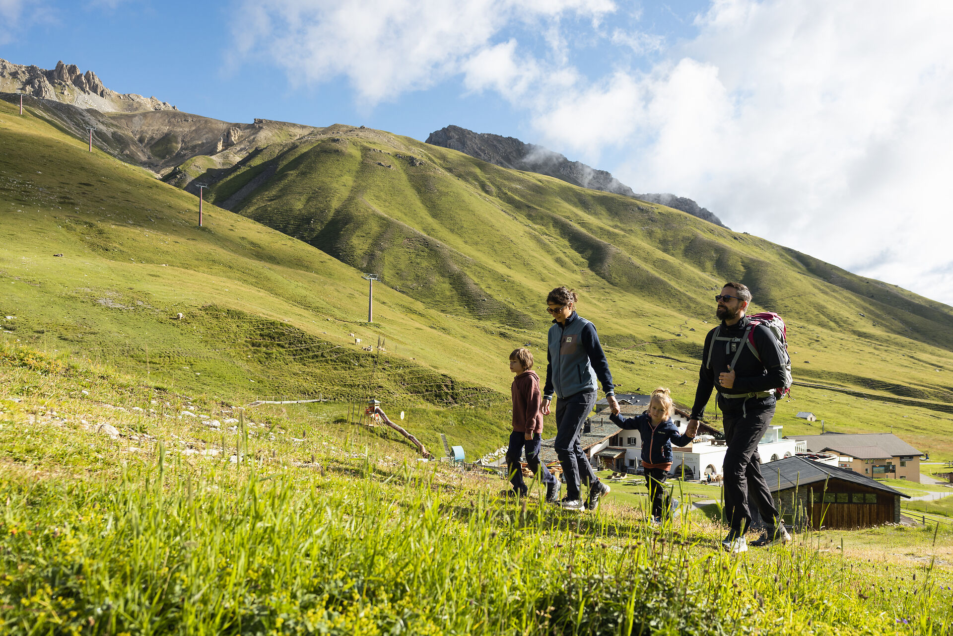 Eine vierköpfige Familie gemeinsam am Wandern auf Marguns. Im Hintergrund ist die Bergstation Marguns und die grüne Berglandschaft des Engadins.