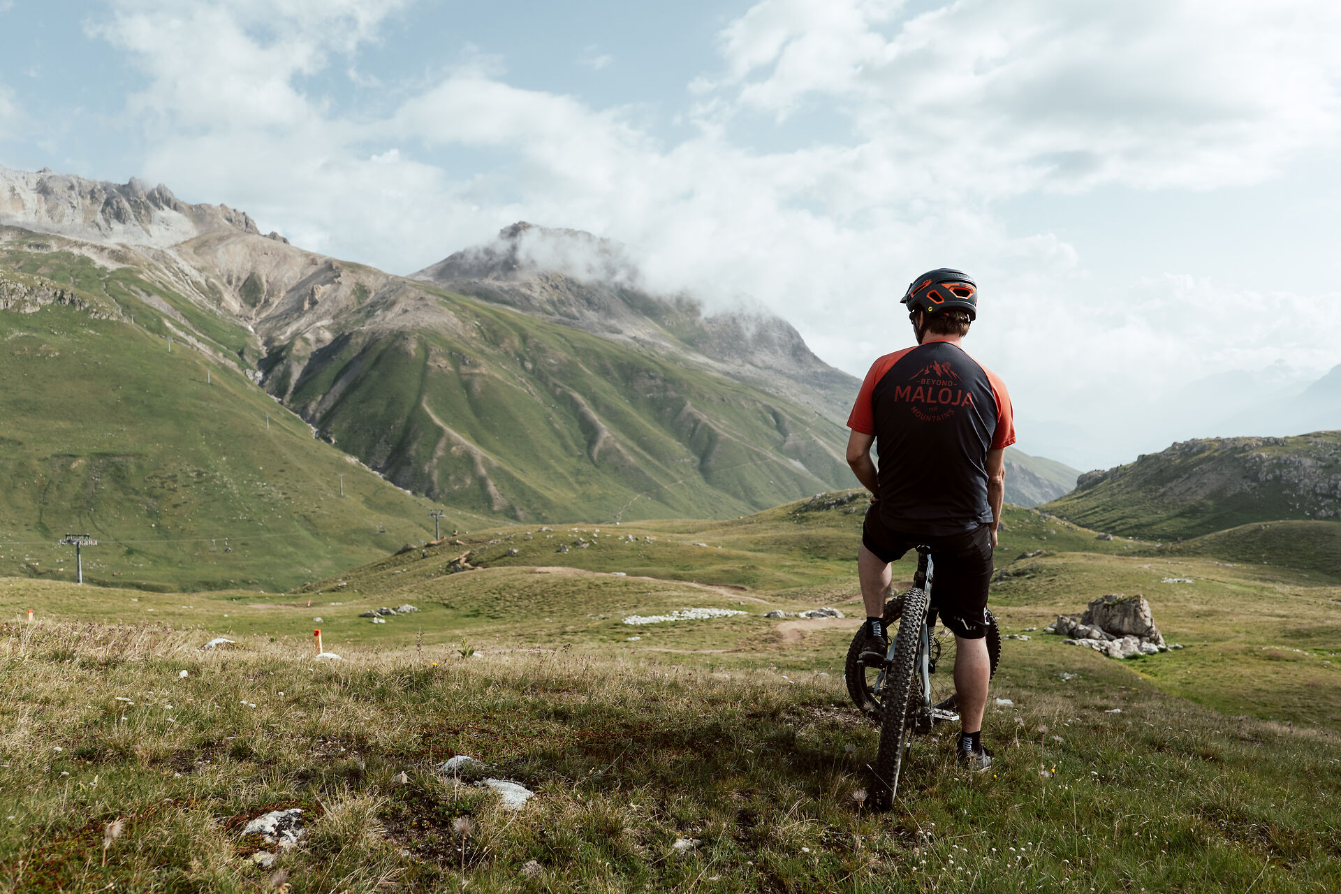 Ein in schwarz-roter Montur gekleideter Mountainbiker sitzt auf seinem Bike und schaut auf die grüne Berglandschaft von Corviglia.