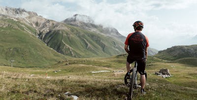 Mountainbike | Corviglia | Sommer Ein in schwarz-roter Montur gekleideter Mountainbiker sitzt auf seinem Bike und schaut auf die grüne Berglandschaft von Corviglia.