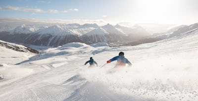 Skifahren Corviglia | Winter Zwei Skifahrer in blau-schwarzer und blau-orangener Skibekleidung fährt auf einer frisch präparierten, breiten Skipiste einen Schwung. Hinter ihm ist aufgewirbelter Schnee zu sehen. Im Hintergrund erstreckt sich eine verschneite Berglandschaft mit klare sich ins Engadiner Tal.
