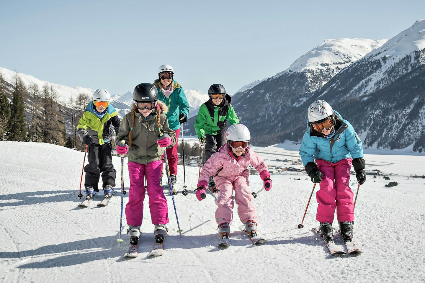 Familienangebote | Corviglia | Winter Fünf Kinder und Jugendliche und eine Mutter fahren Ski. Sie tragen farbige Winterbekleidung und Skibrillen beziehungsweise Helme. Im Hintergrund ist eine weitläufige verschneite Berglandschaft mit bewaldeten Hängen und mehreren Berggipfeln zu sehen.