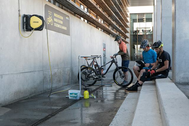 Drei Personen in Fahrradbekleidung mit Helmen bei der Reinigung von Mountainbikes vor dem Gebäude der INN LODGE. Ein Mountainbike steht an einer Halterung und wird mit einem Wasserschlauch gereinigt, während zwei weitere Personen auf einer Treppe sitzen. Neben den Fahrrädern stehen Reinigungsmittel.
