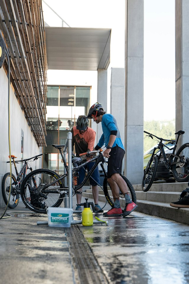 Drei Personen in Fahrradbekleidung mit Helmen bei der Reinigung von Mountainbikes vor dem Gebäude der INN LODGE. Ein Mountainbike steht an einer Halterung und wird mit einem Wasserschlauch gereinigt, während zwei weitere Personen auf einer Treppe sitzen. Neben den Fahrrädern stehen Reinigungsmittel und eine Trinkflasche.