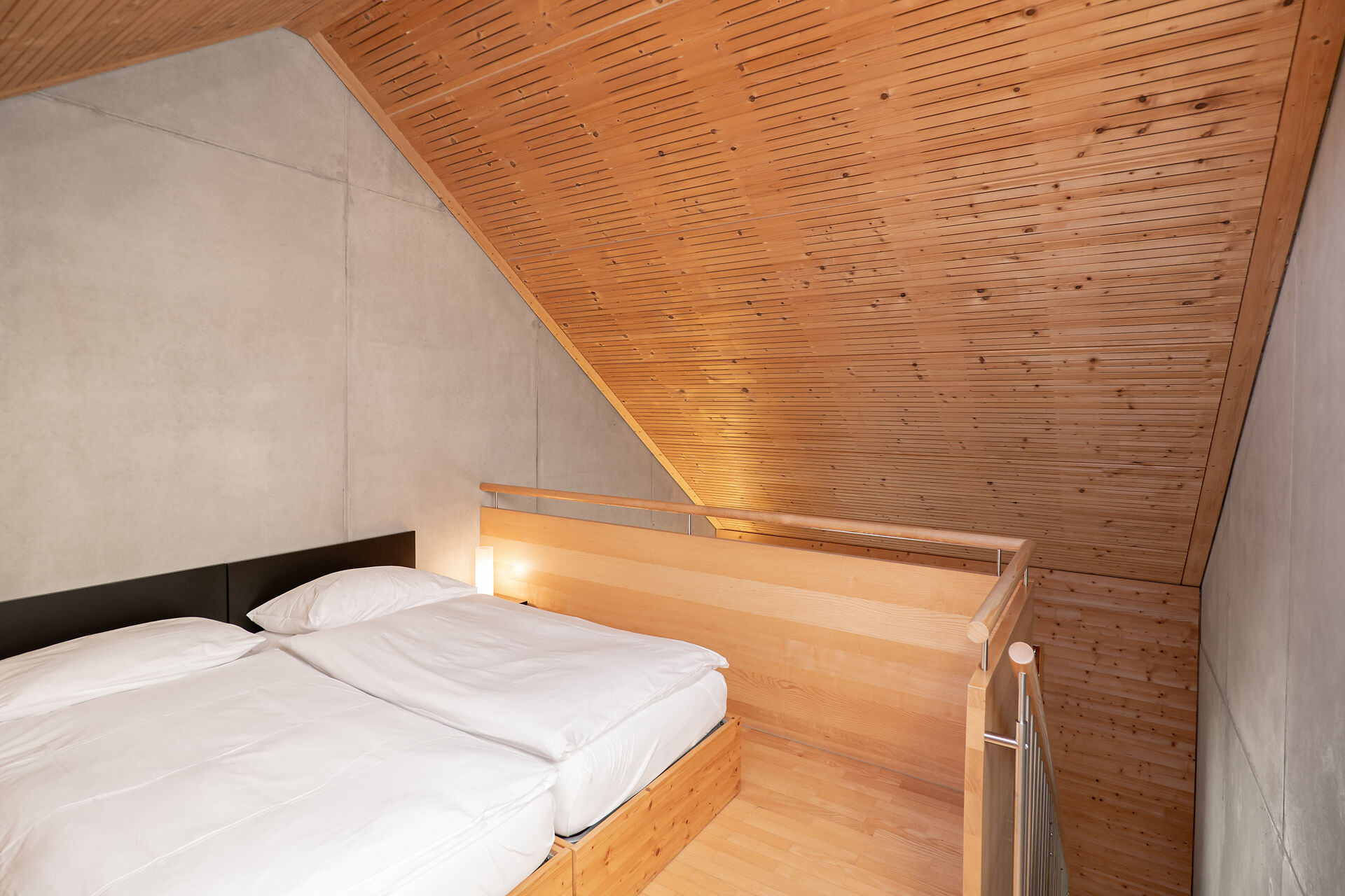 Schlafbereich eines Studio M der Inn Lodge mit zwei zusammengestellten Betten in einem Holzrahmen und weisser Bettwäsche. Neben dem Bett befindet sich eine kleine Leuchte. Der Schlafbereich liegt unter einer grossen, schrägen Holzdecke und ist durch ein Holzgeländer mit Metallstäben vom darunterliegenden Bereich abgetrennt. Die Wände sind teilweise aus hellem Beton und Holz gestaltet.