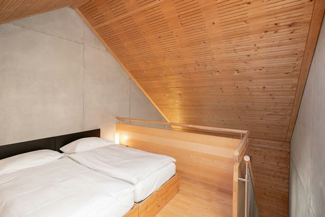 Schlafbereich eines Studio M der Inn Lodge mit zwei zusammengestellten Betten in einem Holzrahmen und weisser Bettwäsche. Neben dem Bett befindet sich eine kleine Leuchte. Der Schlafbereich liegt unter einer grossen, schrägen Holzdecke und ist durch ein Holzgeländer mit Metallstäben vom darunterliegenden Bereich abgetrennt. Die Wände sind teilweise aus hellem Beton und Holz gestaltet.