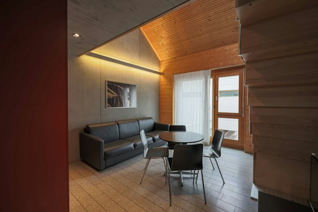 Wohnbereich eines Studio M der Inn Lodge mit schwarzem Ledersofa und rundem Tisch mit vier schwarzen Stühlen. An der grauen Wand hinter dem Sofa hängt ein gerahmtes Bild. Der Raum verfügt über eine teilweise holzverkleidete Decke und Wand sowie ein grosses Fenster und eine Glastür mit weissen Vorhängen. Rechts ist eine holzverkleidete Treppe zu sehen.