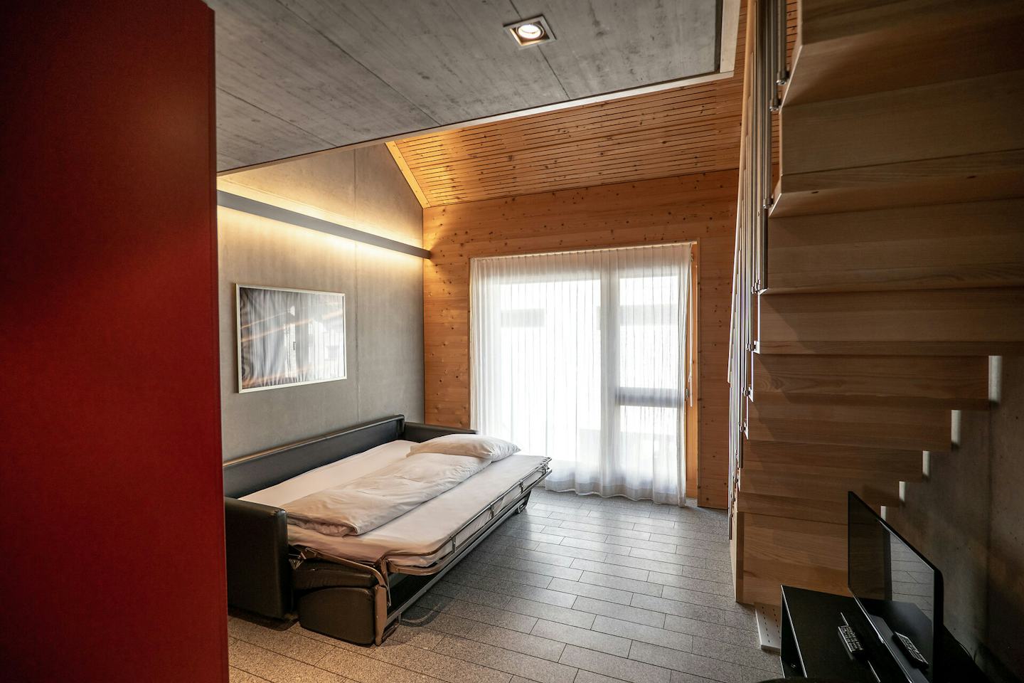 Studio M | ALL IN ONE HOTEL INN LODGE Wohnbereich eines Studio M der Inn Lodge mit schwarzem Ledersofa als Bett umfunktionert. An der grauen Wand hinter dem Sofa hängt ein gerahmtes Bild. Der Raum verfügt über eine teilweise holzverkleidete Decke und Wand sowie ein grosses Fenster und eine Glastür mit weissen Vorhängen. Rechts ist eine holzverkleidete Treppe zu sehen.