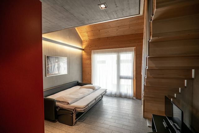 Wohnbereich eines Studio M der Inn Lodge mit schwarzem Ledersofa als Bett umfunktionert. An der grauen Wand hinter dem Sofa hängt ein gerahmtes Bild. Der Raum verfügt über eine teilweise holzverkleidete Decke und Wand sowie ein grosses Fenster und eine Glastür mit weissen Vorhängen. Rechts ist eine holzverkleidete Treppe zu sehen.