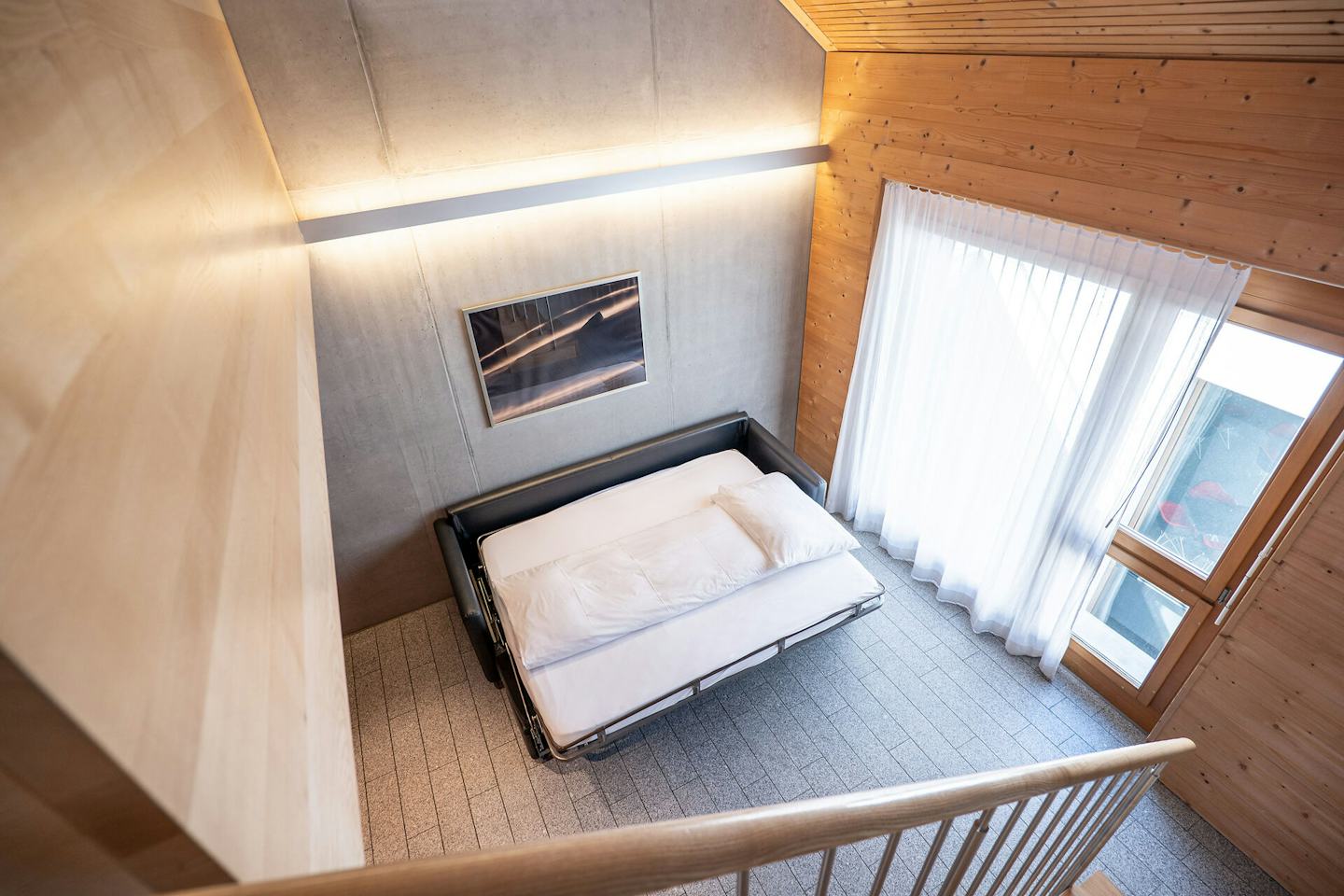 Wohnbereich eines Studio M der Inn Lodge mit einem ausgeklappten schwarzen Schlafsofa und weisser Bettwäsche. Das Schlafsofa steht vor einer grauen Betonwand, an der ein gerahmtes Bild hängt. Rechts befindet sich ein grosses Fenster mit weissen Vorhängen und einer verglasten Tür. Der Raum ist teilweise mit Holz verkleidet und hat einen grauen Boden. Im Vordergrund ist ein Holzgeländer mit Metallstäben zu sehen.