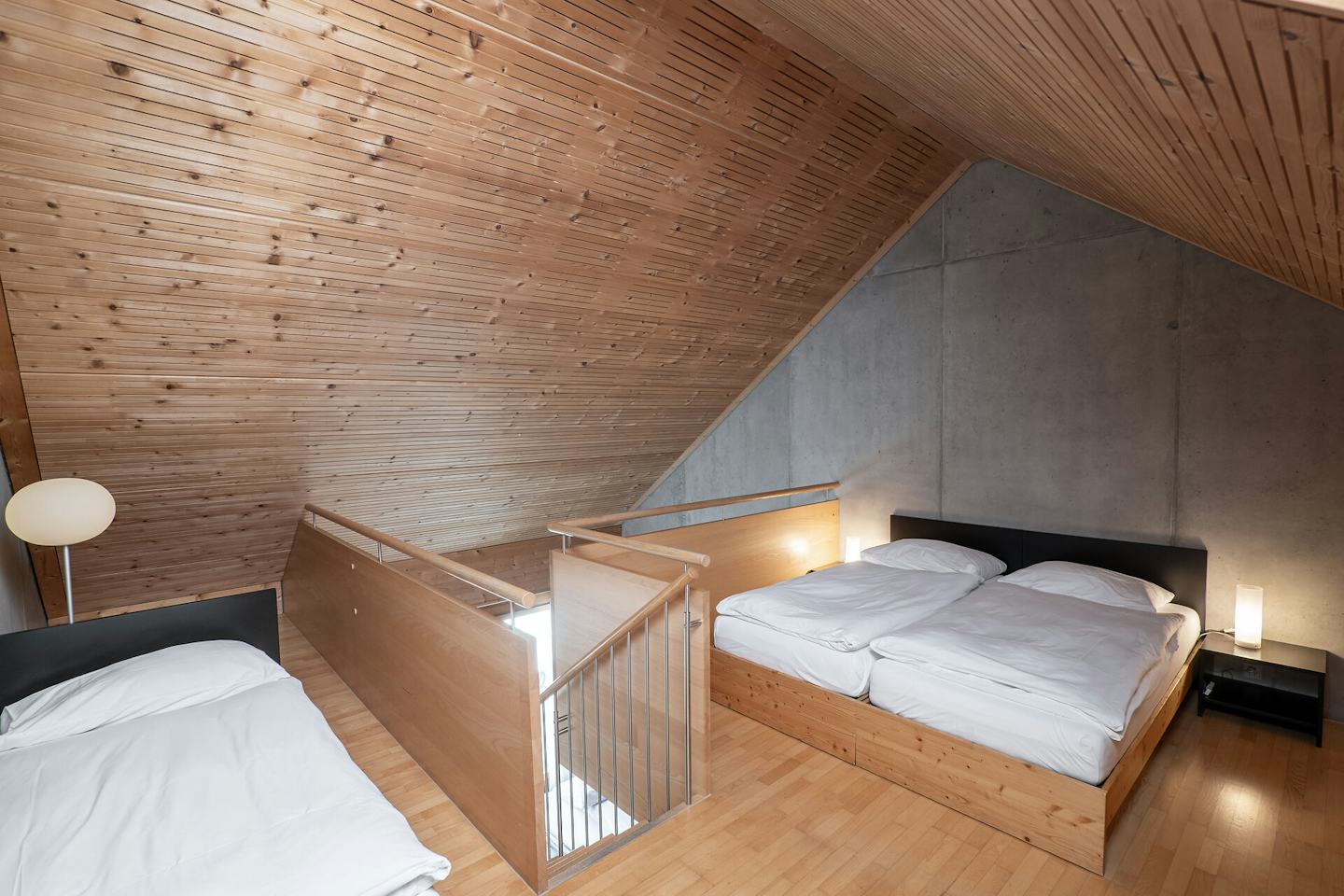 Studio L | ALL IN ONE HOTEL INN LODGE Schlafbereich eines Studio L der Inn Lodge mit zwei Betten auf einer erhöhten Holzplattform und einem weiteren Einzelbett im Vordergrund. Die Betten sind mit weisser Bettwäsche bezogen. Der Raum hat eine Holzdecke und einen Holzboden sowie eine graue Betonwand. Zwischen den Schlafbereichen befindet sich ein Holzgeländer mit Metallstäben, das die Treppe abgrenzt. Neben dem Doppelbett steht ein schwarzer Nachttisch mit einer Leuchte