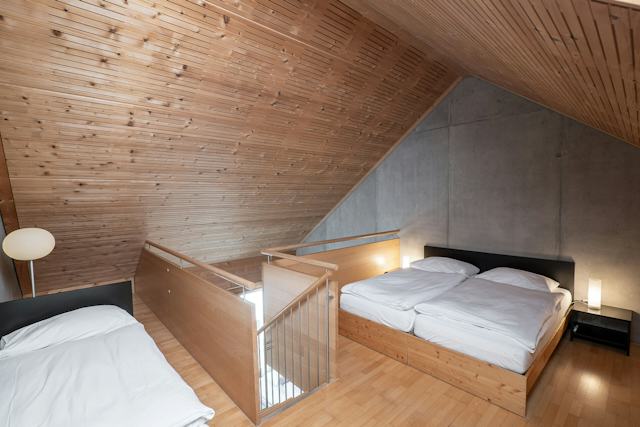Schlafbereich eines Studio L der Inn Lodge mit zwei Betten auf einer erhöhten Holzplattform und einem weiteren Einzelbett im Vordergrund. Die Betten sind mit weisser Bettwäsche bezogen. Der Raum hat eine Holzdecke und einen Holzboden sowie eine graue Betonwand. Zwischen den Schlafbereichen befindet sich ein Holzgeländer mit Metallstäben, das die Treppe abgrenzt. Neben dem Doppelbett steht ein schwarzer Nachttisch mit einer Leuchte