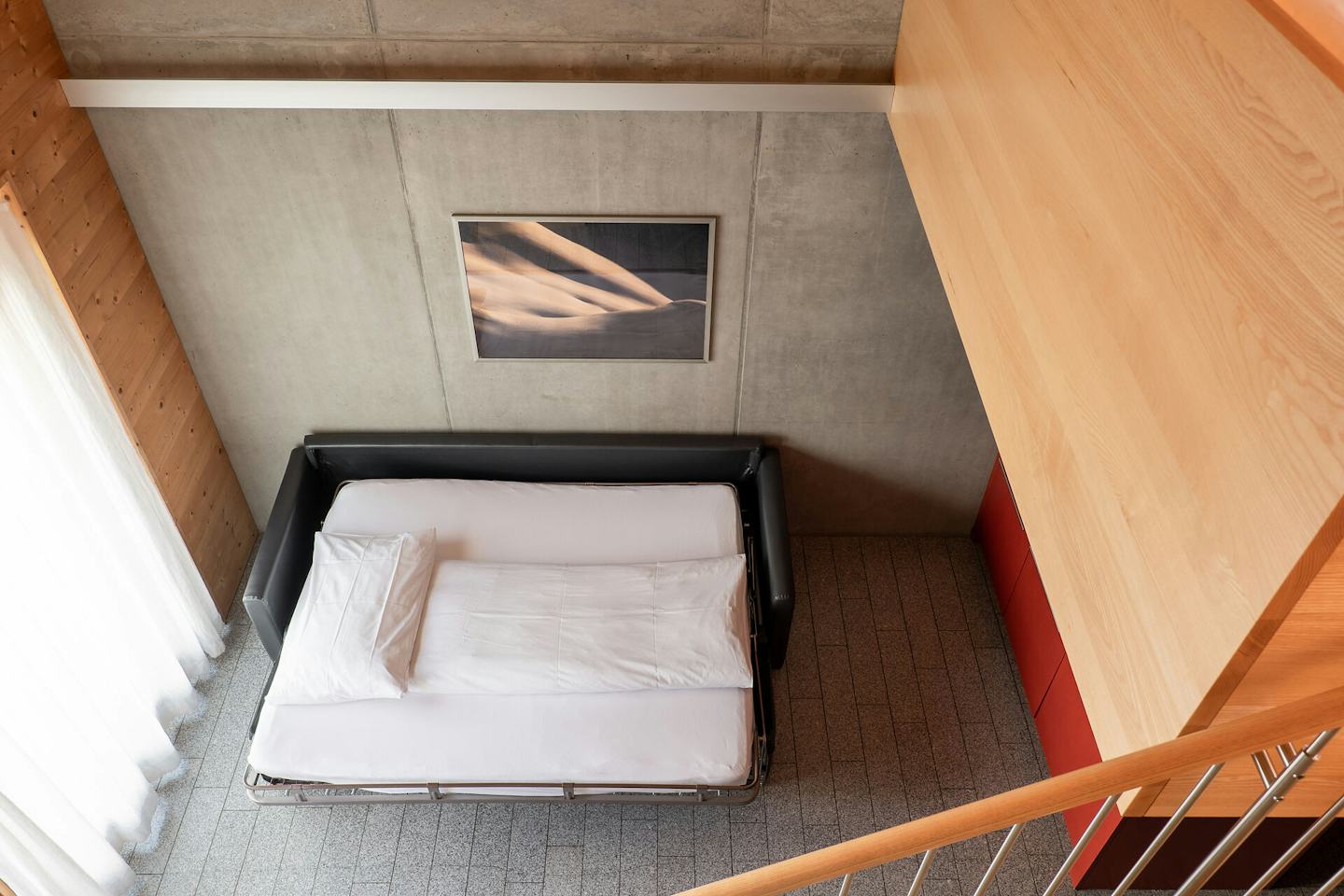 Studio L | ALL IN ONE HOTEL INN LODGE Wohnbereich eines Studio L der Inn Lodge mit einem ausgeklappten schwarzen Schlafsofa und weisser Bettwäsche. Links befindet sich ein grosses Fenster mit weissem Vorhang. Die Wände und Decke sind teilweise aus Holz und grauem Beton gestaltet. An der Betonwand hängt ein gerahmtes Bild. Rechts sind eine rote Küchenfront und eine Holztreppe mit Geländer zu sehen.