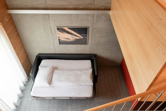 Wohnbereich eines Studio L der Inn Lodge mit einem ausgeklappten schwarzen Schlafsofa und weisser Bettwäsche. Links befindet sich ein grosses Fenster mit weissem Vorhang. Die Wände und Decke sind teilweise aus Holz und grauem Beton gestaltet. An der Betonwand hängt ein gerahmtes Bild. Rechts sind eine rote Küchenfront und eine Holztreppe mit Geländer zu sehen.