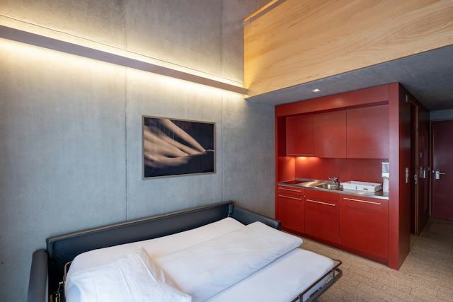 Studio L in der Inn Lodge mit einem ausgeklappten schwarzen Schlafsofa und weisser Bettwäsche vor einer grauen Betonwand. Darüber hängt ein gerahmtes Bild. Rechts befindet sich eine rote Einbauküche mit Spüle, Kochfeld, Schränken und Arbeitsfläche. Im Hintergrund sind eine Tür und ein Eingangsbereich zu sehen.