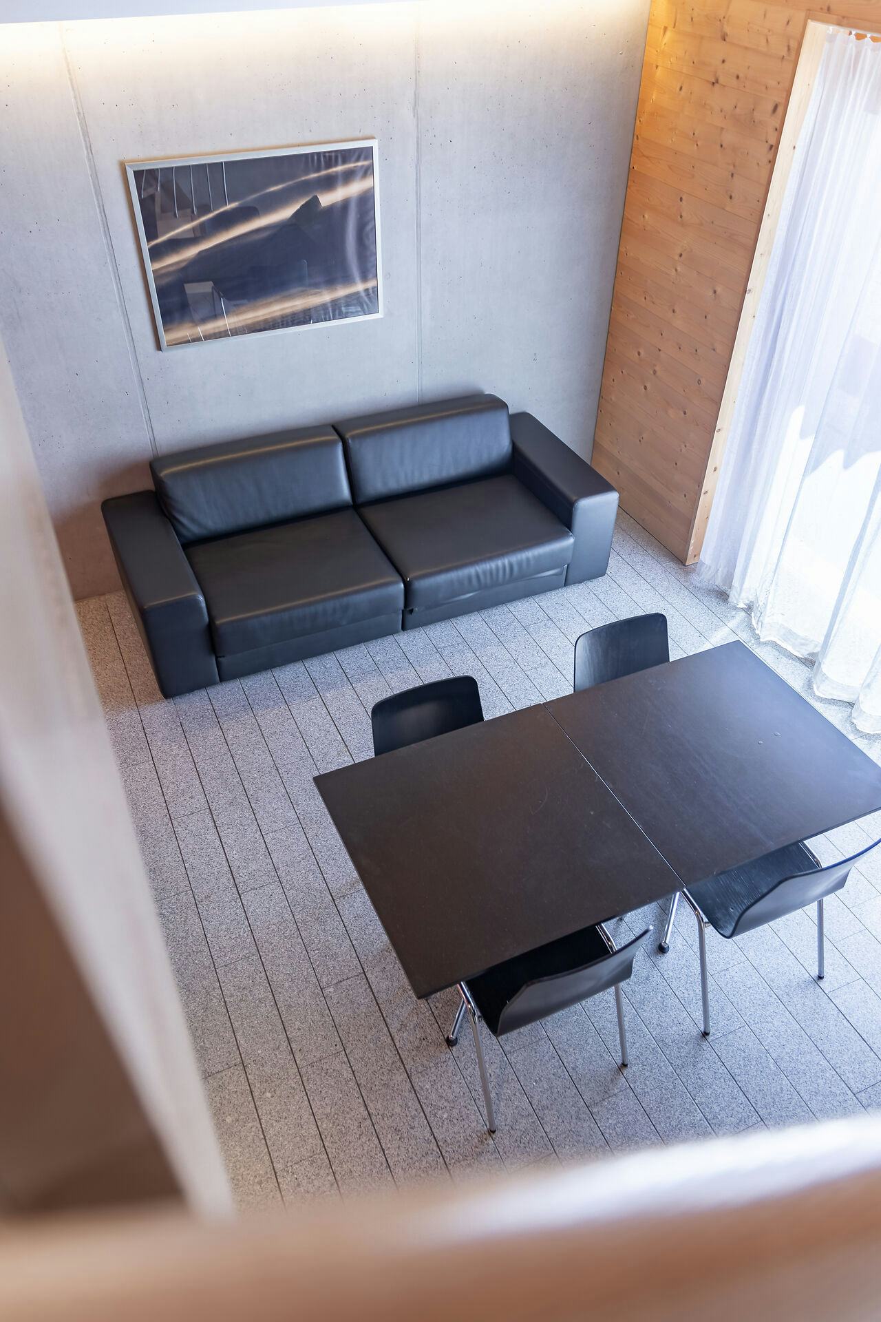Studio XL | ALL IN ONE HOTEL INN LODGE Wohnbereich eines Studio XL der Inn Lodge mit schwarzem Ledersofa und schwarzem Esstisch mit mehreren Stühlen. An der Wand über dem Sofa hängt ein gerahmtes Bild. Der Raum ist teilweise mit Holz verkleidet und verfügt über ein grosses Fenster mit weissen Vorhängen