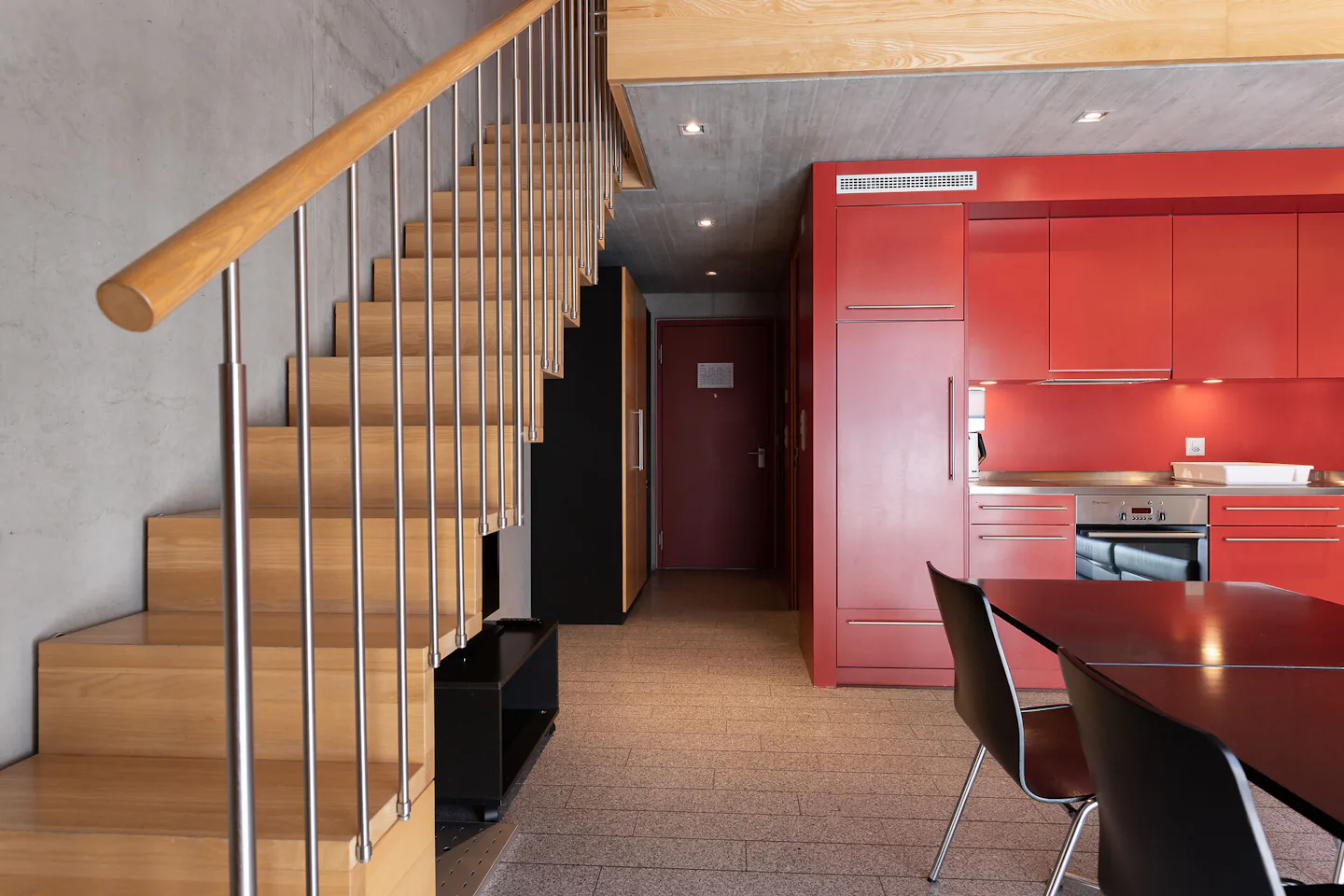 Studio XL | ALL IN ONE HOTEL INN LODGE Innenansicht eines Studio XL der Inn Lodge mit roter Einbauküche, Esstisch und schwarzen Stühlen. Links führt eine Holztreppe mit Metallgeländer in die obere Etage. Im hinteren Bereich sind eine Eingangstür und ein dunkler Einbauschrank zu sehen.