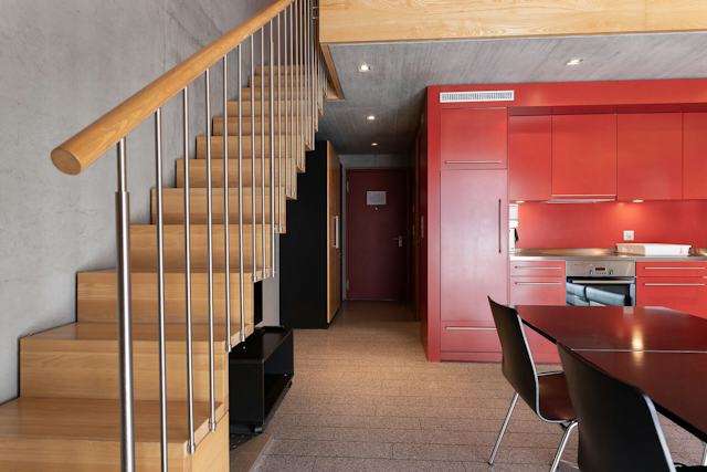 Innenansicht eines Studio XL der Inn Lodge mit roter Einbauküche, Esstisch und schwarzen Stühlen. Links führt eine Holztreppe mit Metallgeländer in die obere Etage. Im hinteren Bereich sind eine Eingangstür und ein dunkler Einbauschrank zu sehen.