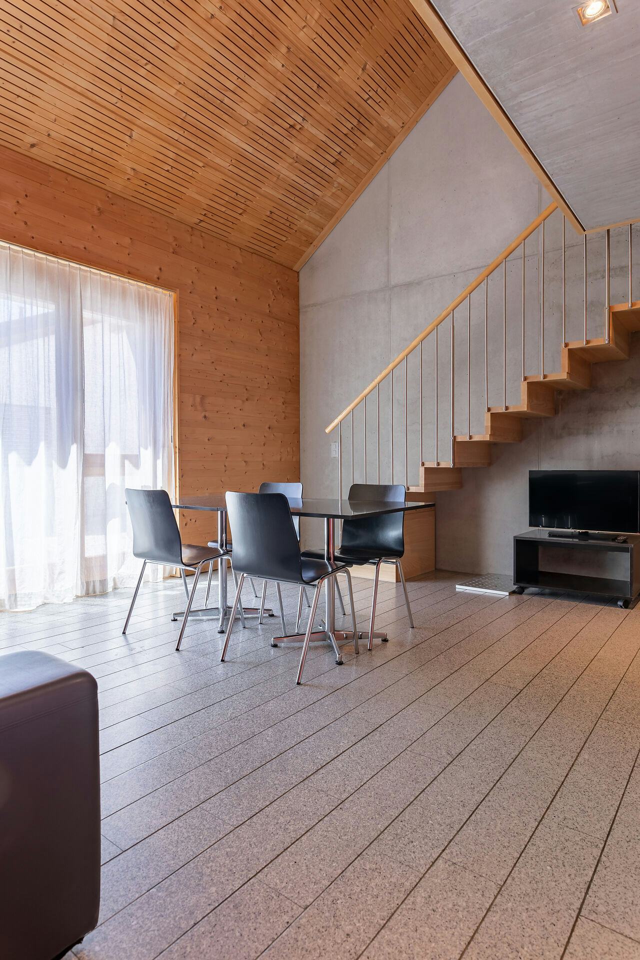 Studio XL | ALL IN ONE HOTEL INN LODGE Wohnbereich eines Studio XL der Inn Lodge mit einem schwarzen Esstisch und vier schwarzen Stühlen. Links ist ein grosses Fenster mit hellen Vorhängen zu sehen. An der Wand rechts befindet sich ein Fernseher auf einem niedrigen Möbelstück. Eine Holztreppe mit Metallgeländer führt vom Wohnbereich in die obere Etage. Der Raum hat einen hellen Boden sowie eine Kombination aus Holz- und Betonwänden.