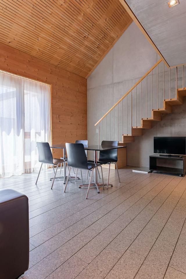 Wohnbereich eines Studio XL der Inn Lodge mit einem schwarzen Esstisch und vier schwarzen Stühlen. Links ist ein grosses Fenster mit hellen Vorhängen zu sehen. An der Wand rechts befindet sich ein Fernseher auf einem niedrigen Möbelstück. Eine Holztreppe mit Metallgeländer führt vom Wohnbereich in die obere Etage. Der Raum hat einen hellen Boden sowie eine Kombination aus Holz- und Betonwänden.
