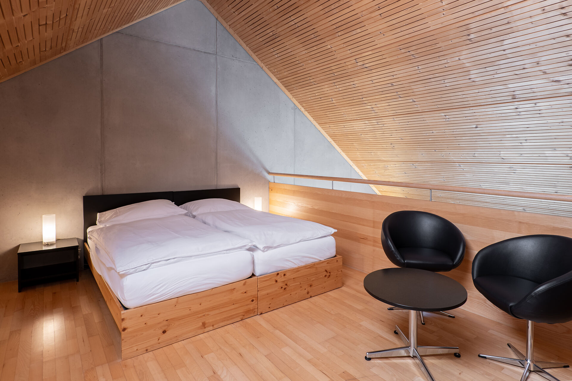 Schlafbereich eines Studio XL der Inn Lodge mit zwei zusammengestellten Betten in einem Holzrahmen. Neben dem Bett steht ein schwarzer Nachttisch mit einer Lampe. Rechts befinden sich zwei schwarze Sessel und ein runder Tisch. Der Raum hat einen Holzboden, eine teilweise holzverkleidete Wand und eine sichtbare Dachschräge.
