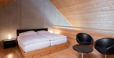 Studio XL | ALL IN ONE HOTEL INN LODGE Schlafbereich eines Studio XL der Inn Lodge mit zwei zusammengestellten Betten in einem Holzrahmen. Neben dem Bett steht ein schwarzer Nachttisch mit einer Lampe. Rechts befinden sich zwei schwarze Sessel und ein runder Tisch. Der Raum hat einen Holzboden, eine teilweise holzverkleidete Wand und eine sichtbare Dachschräge.