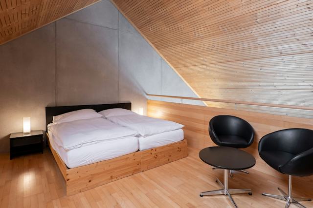 Schlafbereich eines Studio XL der Inn Lodge mit zwei zusammengestellten Betten in einem Holzrahmen. Neben dem Bett steht ein schwarzer Nachttisch mit einer Lampe. Rechts befinden sich zwei schwarze Sessel und ein runder Tisch. Der Raum hat einen Holzboden, eine teilweise holzverkleidete Wand und eine sichtbare Dachschräge.