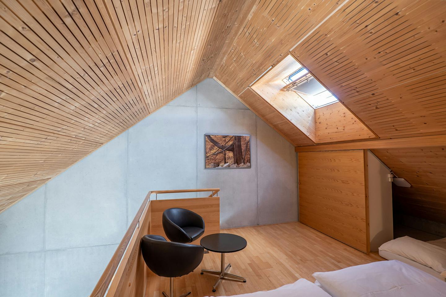 Studio XL | ALL IN ONE HOTEL INN LODGE Schlafbereich eines Studio XL der Inn Lodge. Im Bild steht ein schwarzer Nachttisch mit zwei schwarzen Stühlen. Der Raum hat einen Holzboden, eine teilweise holzverkleidete Wand und eine sichtbare Dachschräge.