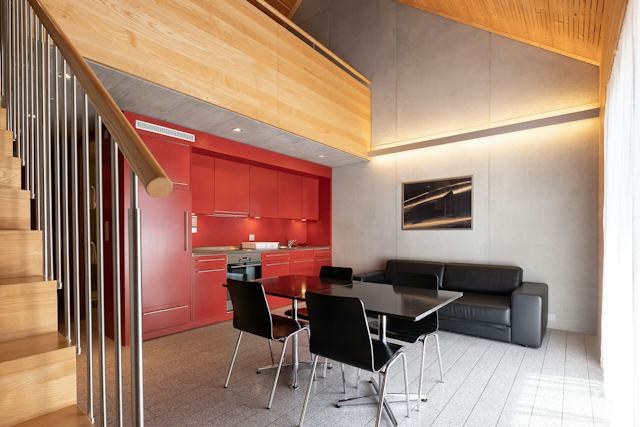 Wohnbereich eines Studio XL der Inn Lodge mit roter Einbauküche, Esstisch und schwarzen Stühlen. Neben dem Essbereich befindet sich eine schwarze Sitzbank, während links eine Holztreppe mit Metallgeländer in die obere Etage führt. An der Wand hängt ein grossformatiges Bild.