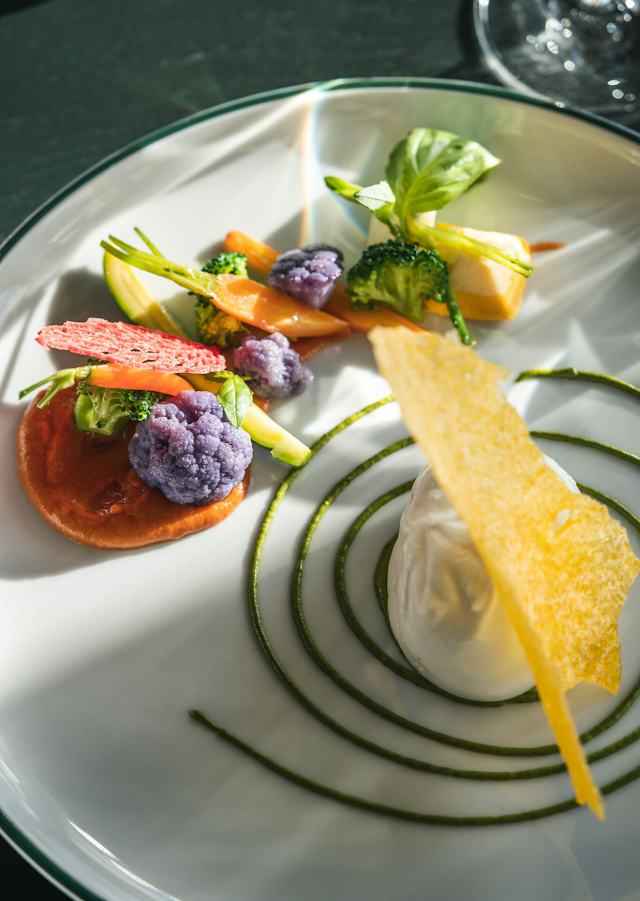 Angerichtete Burrata auf einem weissen Teller mit Tomaten, grünen Blättern und weiteren Zutaten. Das Gericht steht auf einem gedeckten Tisch mit Besteck und Gläsern.