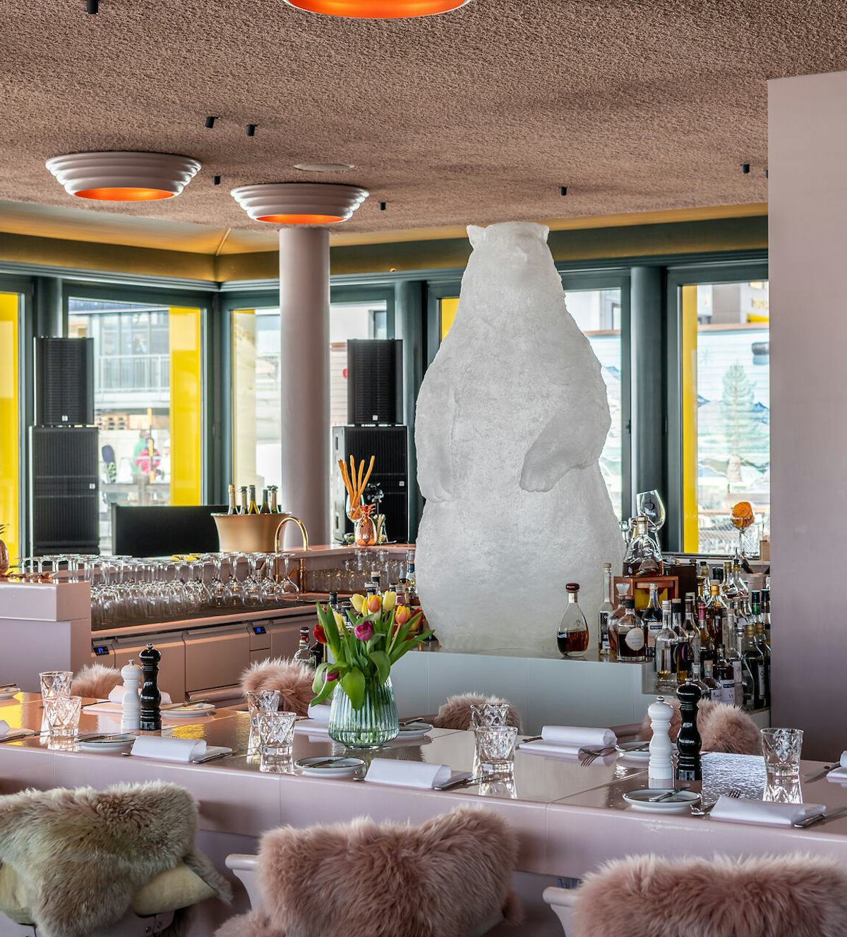Restaurant White Marmot | Corviglia St. Moritz Moderner Innenraum des White Marmot mit einer langen Bar im Mittelpunkt, an der Flaschen, Gläser und verschiedene Dekorationen und ein weisses grosses Murmeltier stehen. Die Bar ist von hellen Stühlen mit Fellbezügen umgeben. Im hinteren Bereich befinden sich weitere Sitzplätze mit grünen Polsterstühlen und Tischen.