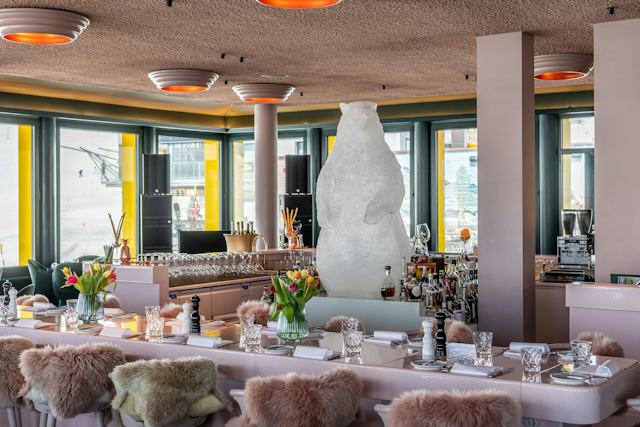 Moderner Innenraum des White Marmot mit einer langen Bar im Mittelpunkt, an der Flaschen, Gläser und verschiedene Dekorationen und ein weisses grosses Murmeltier stehen. Die Bar ist von hellen Stühlen mit Fellbezügen umgeben. Im hinteren Bereich befinden sich weitere Sitzplätze mit grünen Polsterstühlen und Tischen.