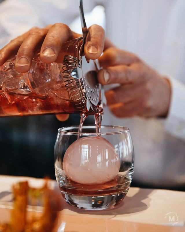 Ein dunkel orangener Cocktail wird über einen runden Eisblock, der sich im Glas befindet, eingeschenkt.
