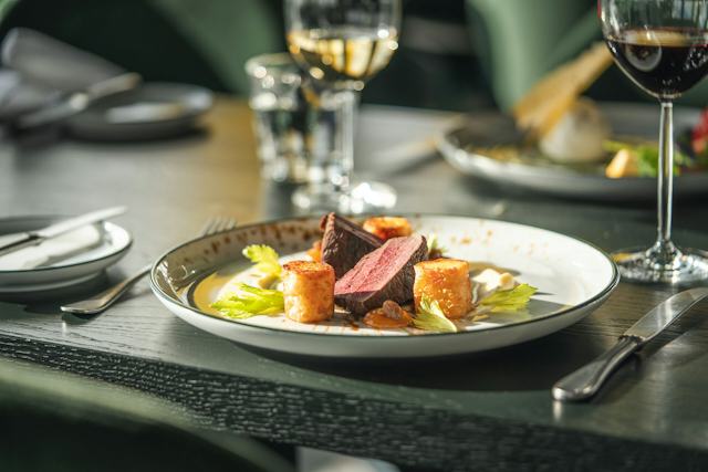 Angerichtetes Hirschentrecôte auf einem weissen Teller mit Beilagen und Sauce. Im Hintergrund sind weitere Teller und Besteck und zwei Weingläser auf einem gedeckten Tisch zu sehen.