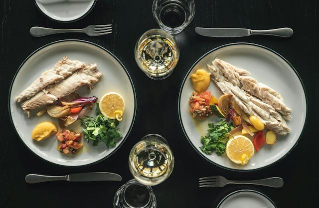 Zwei Teller mit einem aufwendig angerichtetem Gericht mit einem gebratenen Fischfilet, Gemüse und einer knusprigen Beilage auf einem weissen Teller. Daneben steht ein kleiner Teller mit einer weiteren Speise.