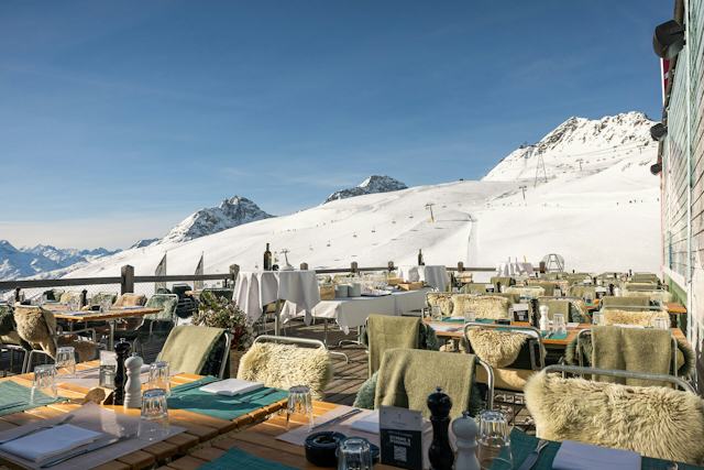 Sonnige Terrasse des White Marmot auf Corviglia mit gedeckten Tischen und Sitzplätzen. Im Hintergrund sind die, Pisten des Skigebiets Vorviglia, die umliegenden Berge und die alpine Landschaft zu sehen.