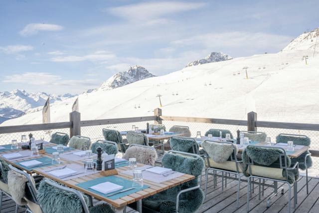 Sonnige Terrasse des White Marmot auf Corviglia mit gedeckten Tischen und Sitzplätzen. Im Hintergrund sind die, Pisten des Skigebiets Corviglia, eine Sesselbahn, die umliegenden Berge und die alpine Landschaft zu sehen.
