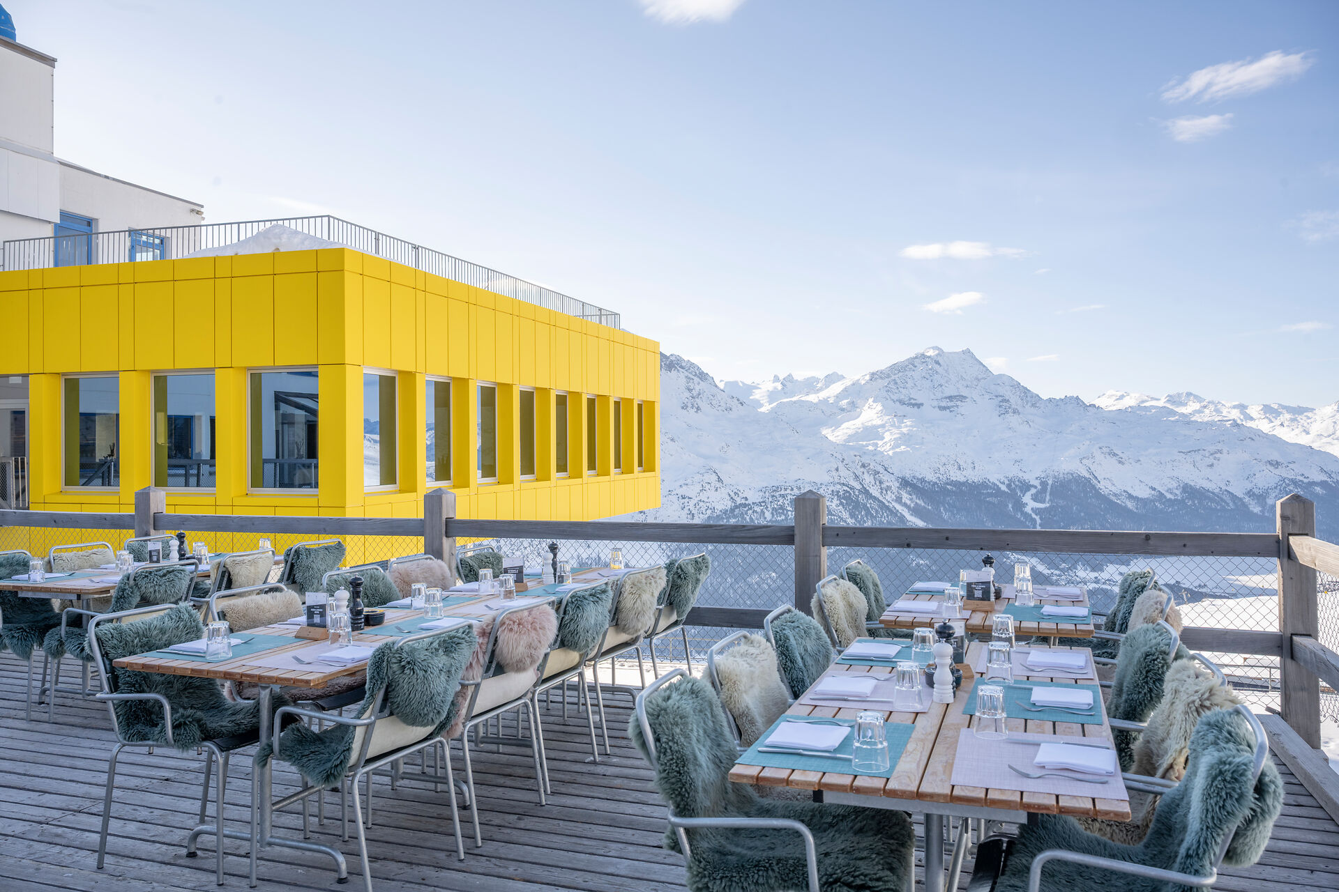 Terrasse des White Marmot auf Corviglia mit Sitzplätzen und Tischen vor dem gelben Gebäudeteil der Bergstation Corviglia und der verschneiten Bergkulisse des Engadins.