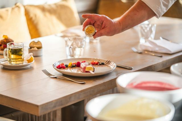 Hand streut Goldstaub über einen angerichteten Teller mit dem Corviglia Magnum und Früchten auf einem gedeckten Tisch im White Marmot. Daneben stehen ein Glas Tee sowie Gläser, Besteck und weitere Teller.