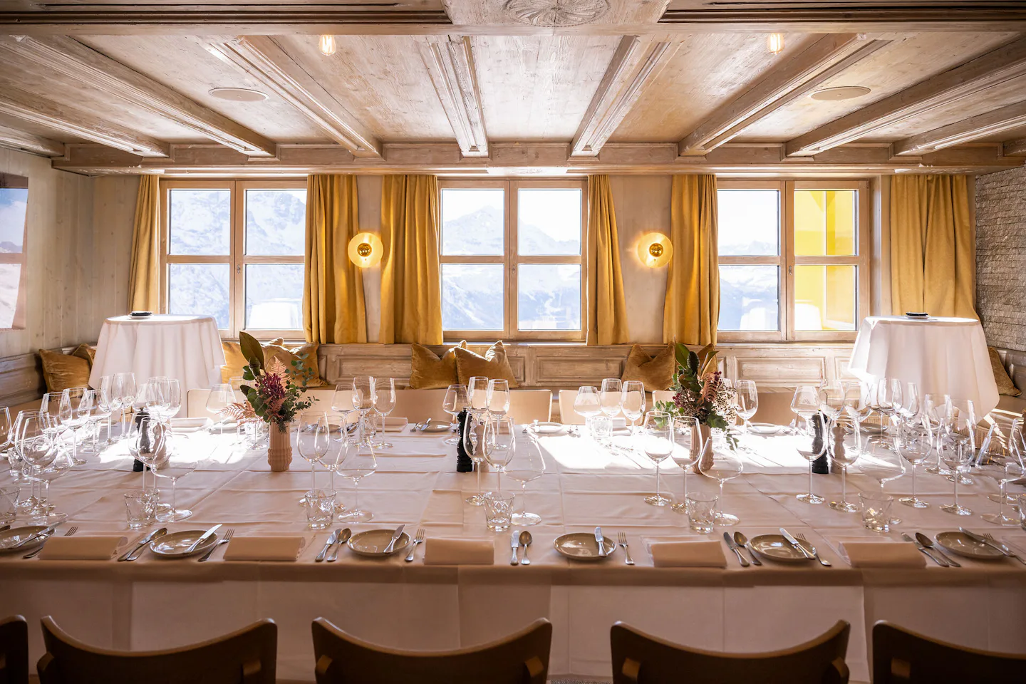 Restaurant White Marmot | Corviglia St. Moritz Festlich gedeckter Tisch im Golden Eye des White Marmot auf Corviglia mit weissen Tischdecken, Gläsern, Besteck und Blumendekoration. Durch die grossen Fenster ist die verschneite Berglandschaft zu sehen, im Raum befinden sich zudem eine Steinwand mit Kamin und eine Holzdecke.