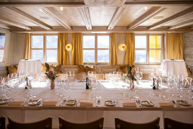 Festlich gedeckter Tisch im Golden Eye des White Marmot auf Corviglia mit weissen Tischdecken, Gläsern, Besteck und Blumendekoration. Durch die grossen Fenster ist die verschneite Berglandschaft zu sehen, im Raum befinden sich zudem eine Steinwand mit Kamin und eine Holzdecke.