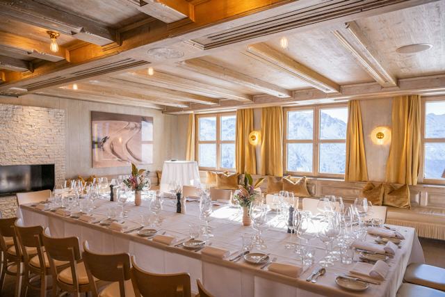 Festlich gedeckter langer Tisch im Golden Eye des White Marmot auf Corviglia mit weissen Tischdecken, zahlreichen Weingläsern, Besteck und floralen Tischdekorationen. Im Hintergrund befinden sich eine Steinwand mit Kamin, eine Holzdecke sowie grosse Fenster mit Blick auf die verschneite Berglandschaft.