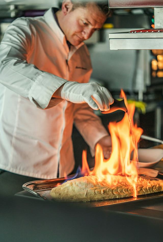White Marmot Koch in weisser Kochjacke und weissen Handschuhen beim Zubereiten eines Gerichts in einer professionellen Küche. Vor ihm ist eine Pfanne mit sichtbarer Flamme zu sehen, während er mit der Hand daran arbeitet. Im Hintergrund befinden sich Küchengeräte und Arbeitsflächen.