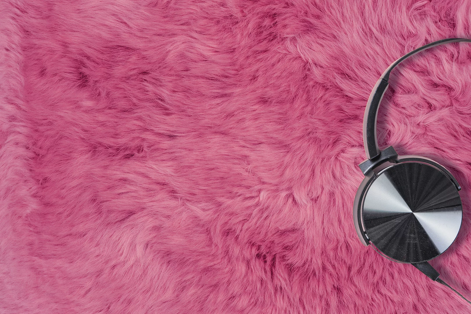 Schwarze Kopfhörer liegen auf einem flauschigen pinkfarbenen Fell. Die Kopfhörer befinden sich auf der rechten Seite des Bildes, während das Fell den gesamten Hintergrund ausfüllt