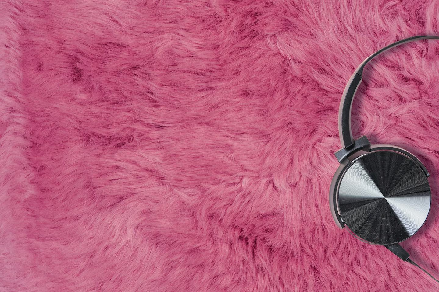 Schwarze Kopfhörer liegen auf einem flauschigen pinkfarbenen Fell. Die Kopfhörer befinden sich auf der rechten Seite des Bildes, während das Fell den gesamten Hintergrund ausfüllt