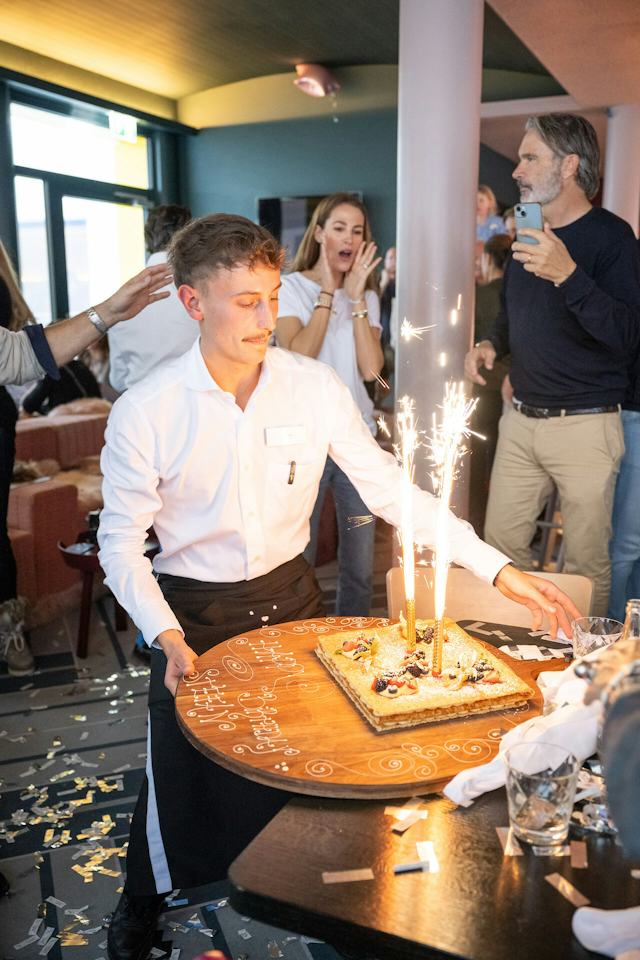 Ein Kellner in weisser Jacke mit einem Tablett bei einem Lunch & Dance -Event im White Marmot. Im Hintergrund stehen zwei Gäste.