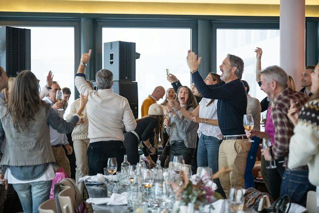 Gäste feiern bei einem Lunch & Dance -Event im White Marmot. Im Vordergrund ist ein gedeckter Tisch. Mehrere Personen halten ebenfalls Gläser mit Getränken in der Hand. Im Hintergrund sitzen und stehen zahlreiche weitere Gäste.