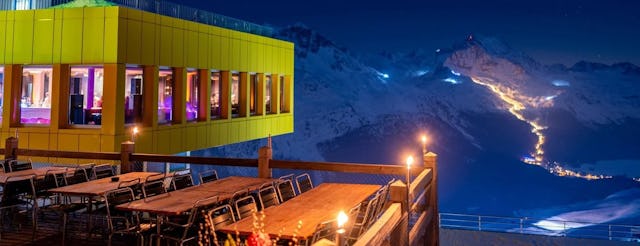 Aussenterrasse des White Marmot bei Nacht mit zahlreichen Holztischen und Metallstühlen. Die Terrasse ist mit warm leuchtenden Laternen entlang des Geländers beleuchtet. Links ist der gelbe Gebäudeteil der Bergstation Corviglia mit grossen Fenstern zu sehen, durch die der farbig beleuchtete Innenraum sichtbar ist. Im Hintergrund erstreckt sich eine nächtliche verschneite Berglandschaft mit beleuchteten Berg- und Talbereichen.