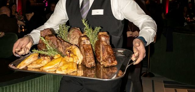 Mitarbeiter eines Restaurants trägt ein grosses Serviertablett mit mehreren gebratenen Fleischstücken und goldbraunen Beilagen. Er trägt ein weisses Hemd, eine schwarze Weste und eine Krawatte. Im Hintergrund sind weitere Gäste an Tischen, dunkle Fensterflächen und eine beleuchtete Innenraumgestaltung mit warmen gelben und roten Lichtakzenten zu sehen.